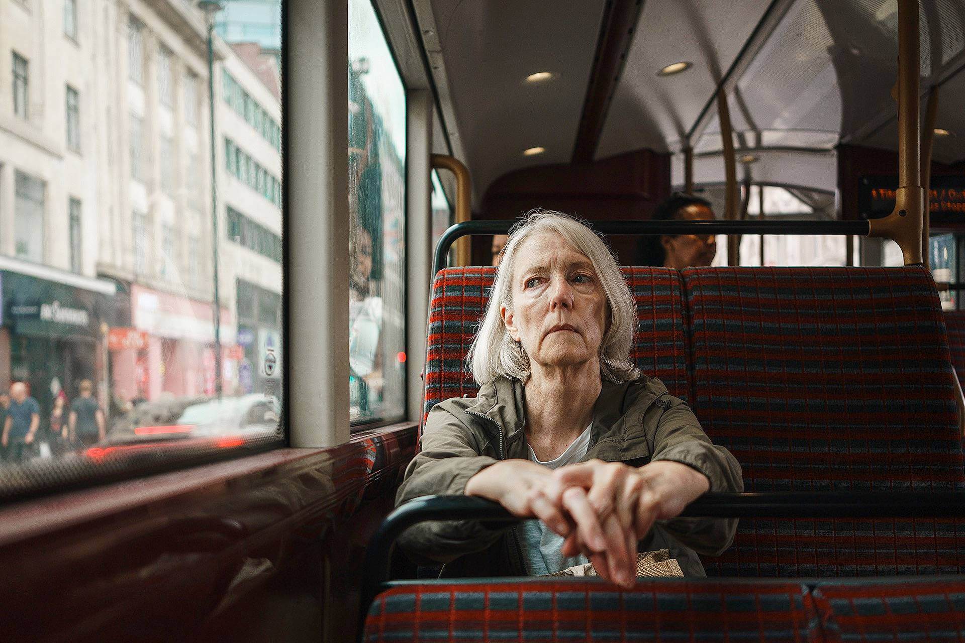 An older woman with gray hair sits alone on a city bus, looking out the window with a neutral expression. Urban buildings and pedestrians are visible outside.