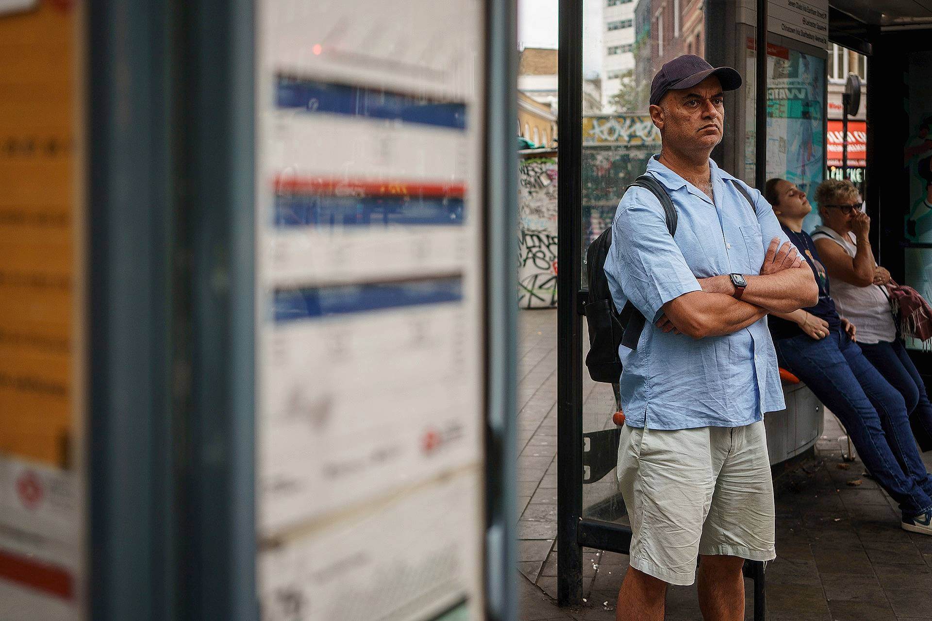 A man in a light blue shirt and shorts stands with arms crossed at a bus stop, while others sit and wait in the background.