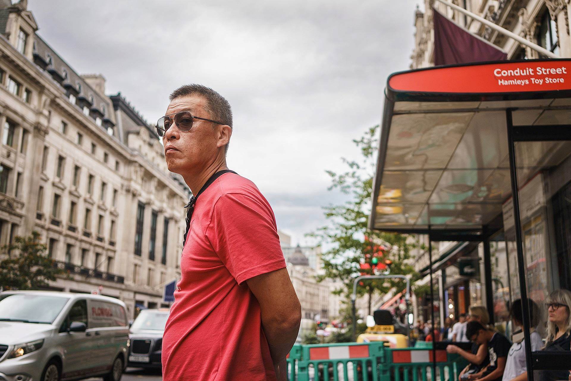 A man in a red shirt and sunglasses stands on a busy city street near a bus stop with cars and buildings in the background.