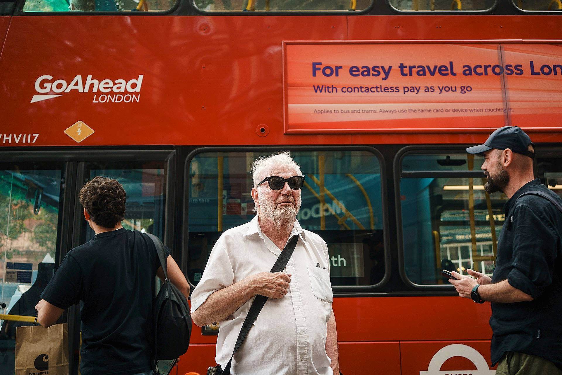 An older man in sunglasses stands in front of a red London double-decker bus, with two other men nearby; the bus displays an ad about contactless payment.