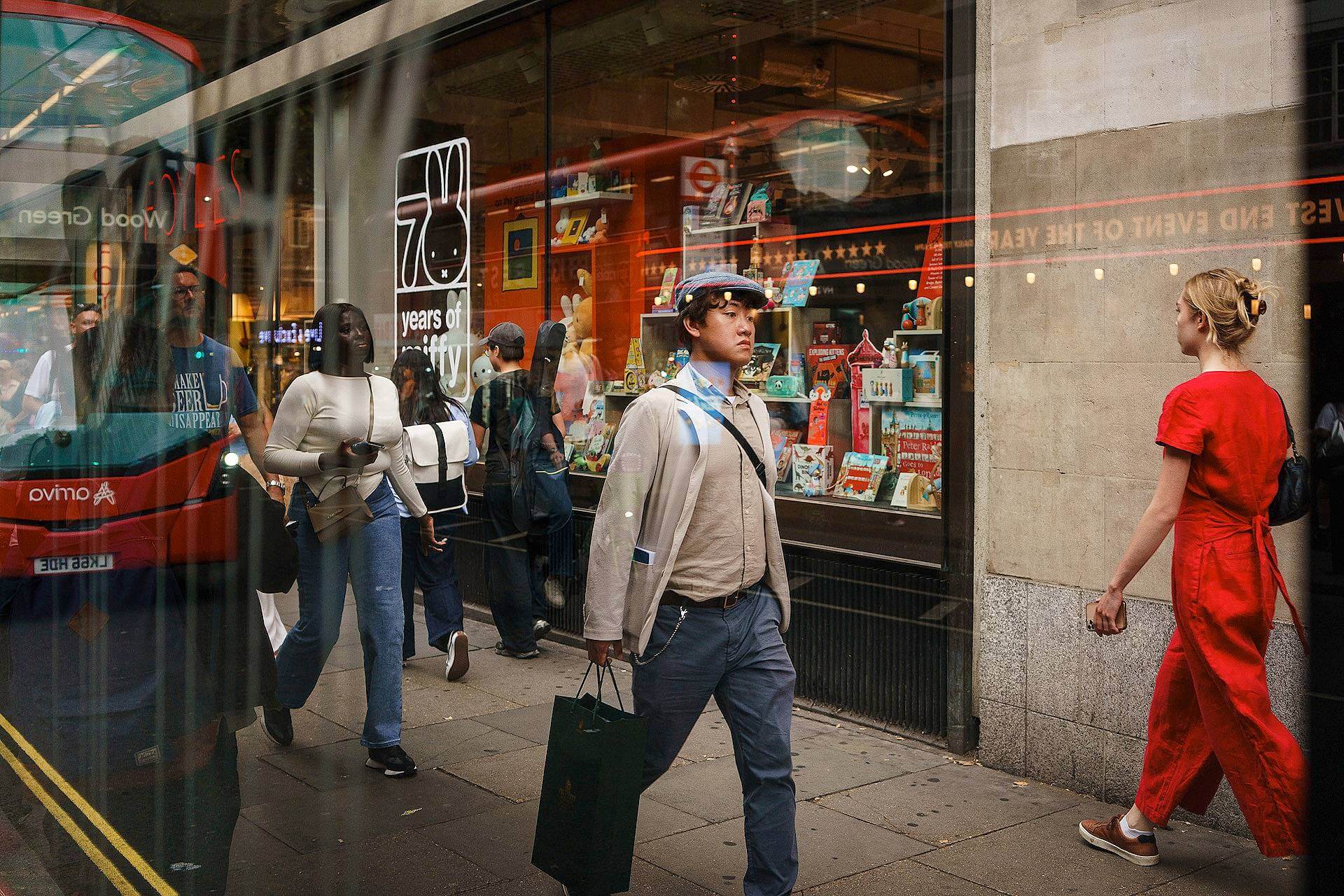 People walk past a bookstore on a city street, with a red bus reflected in the window and colorful books displayed inside the shop.