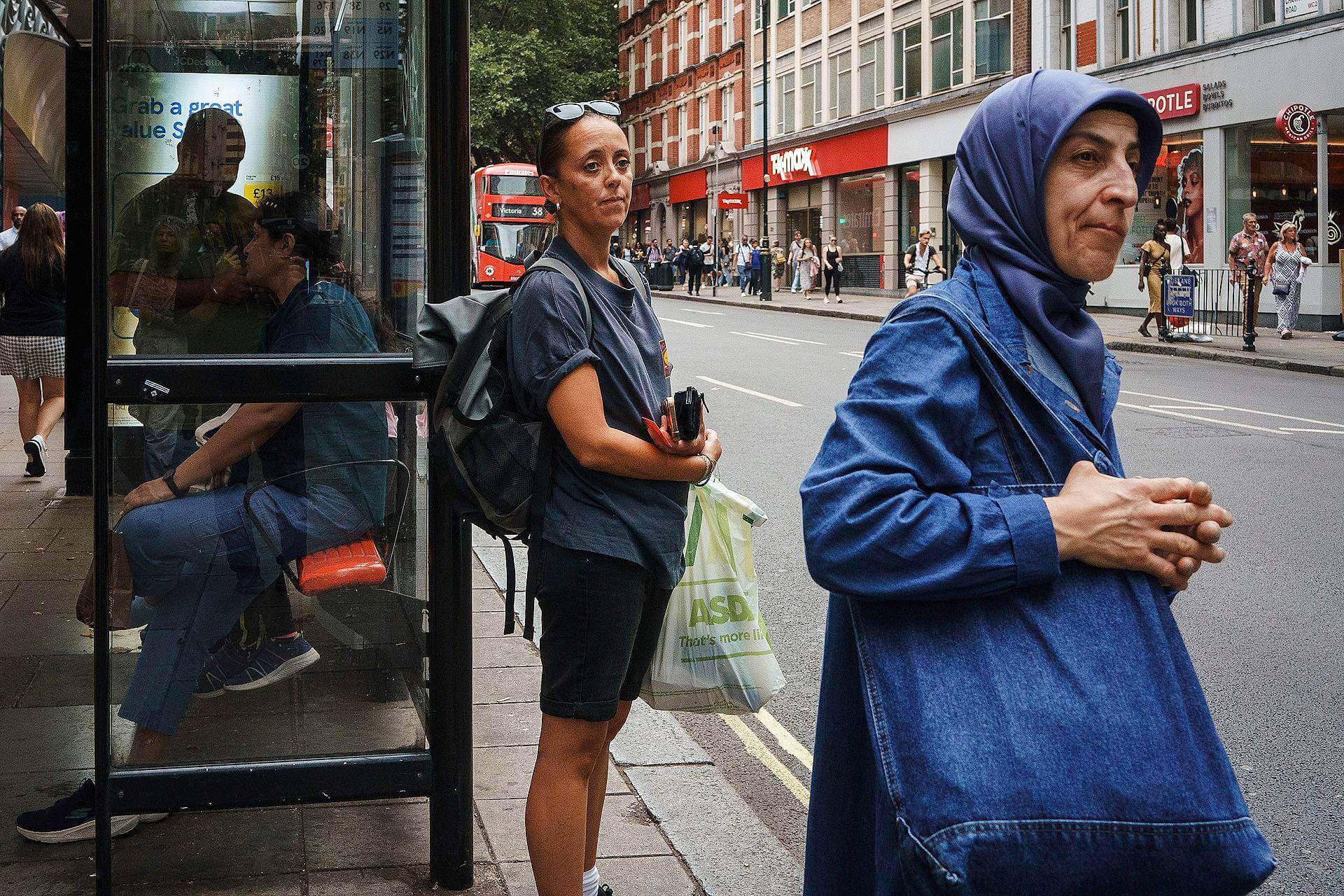 A woman in a denim jacket and headscarf walks past a bus stop where another woman stands holding shopping bags; people and a bus are visible in the background on a busy city street.