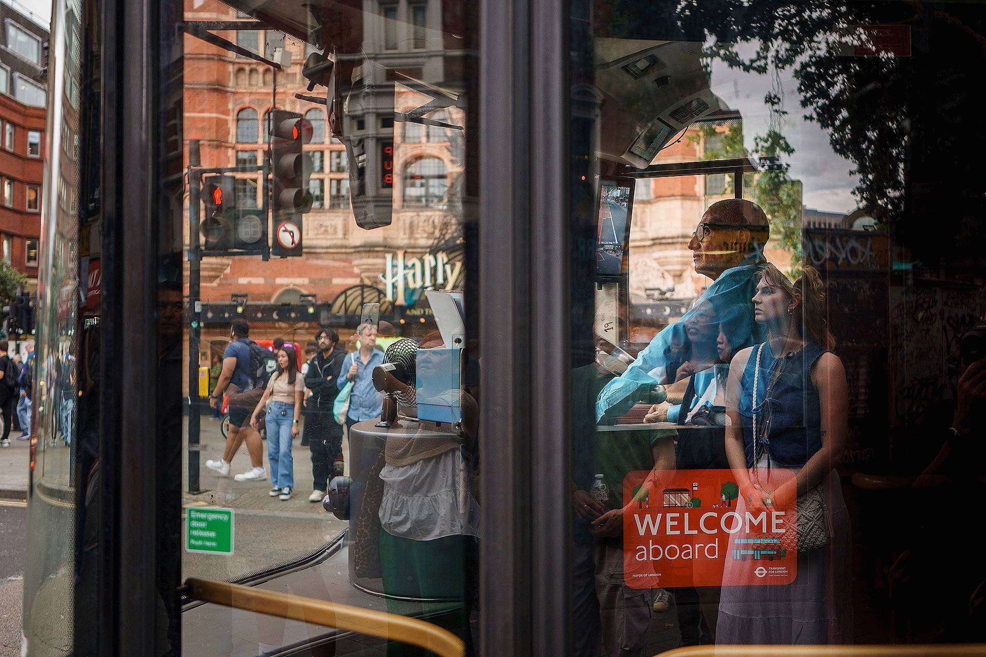 People are reflected in a bus window on a busy city street, with a "Welcome Aboard" sign visible and Harry Potter-themed shop in the background.