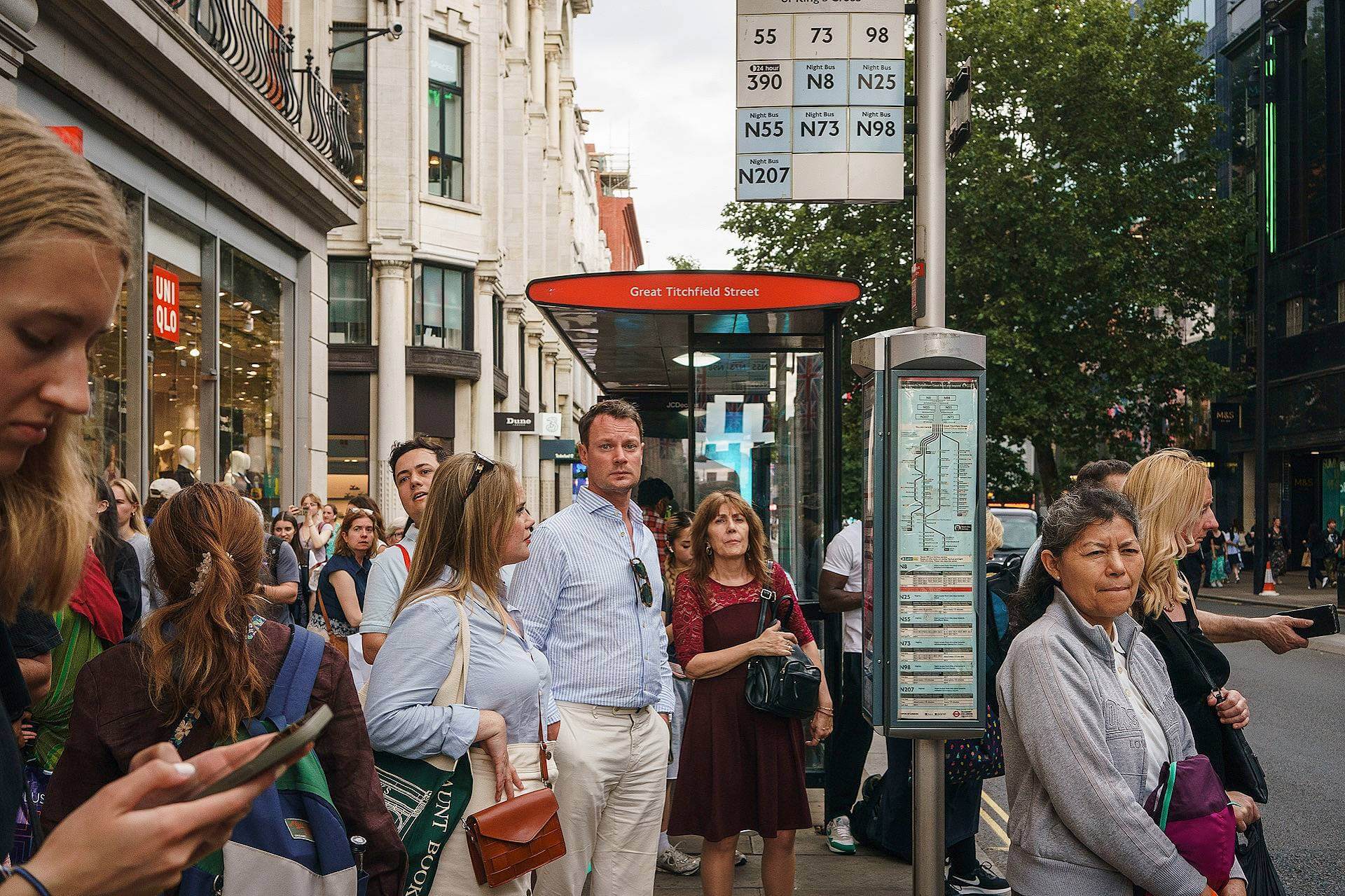 A group of people wait at a busy London bus stop under a sign listing bus routes on Great Titchfield Street; some are looking at phones, others look ahead.