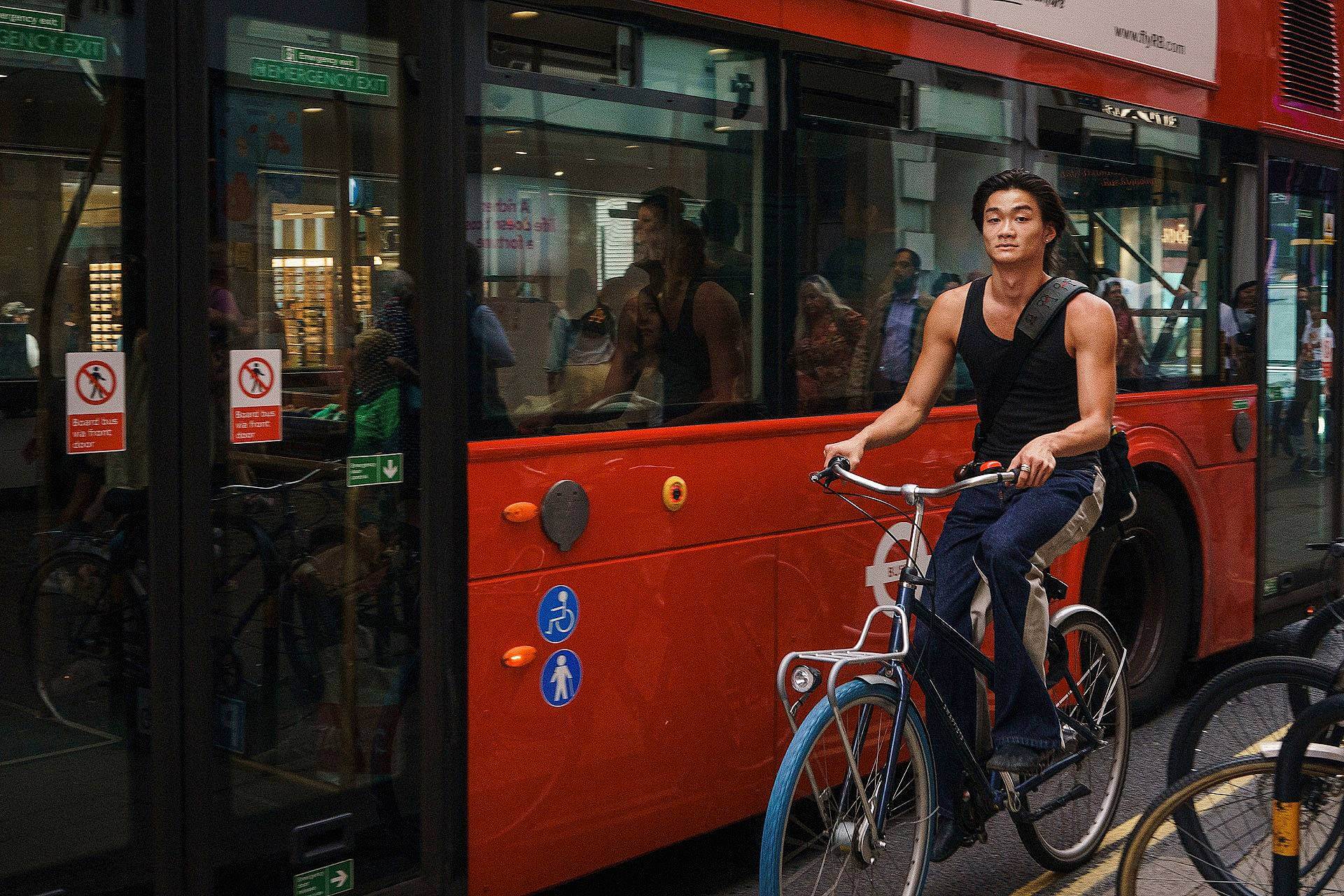 A man rides a bicycle past a red city bus on a busy street, with reflections of people in the bus windows and shops visible in the background.
